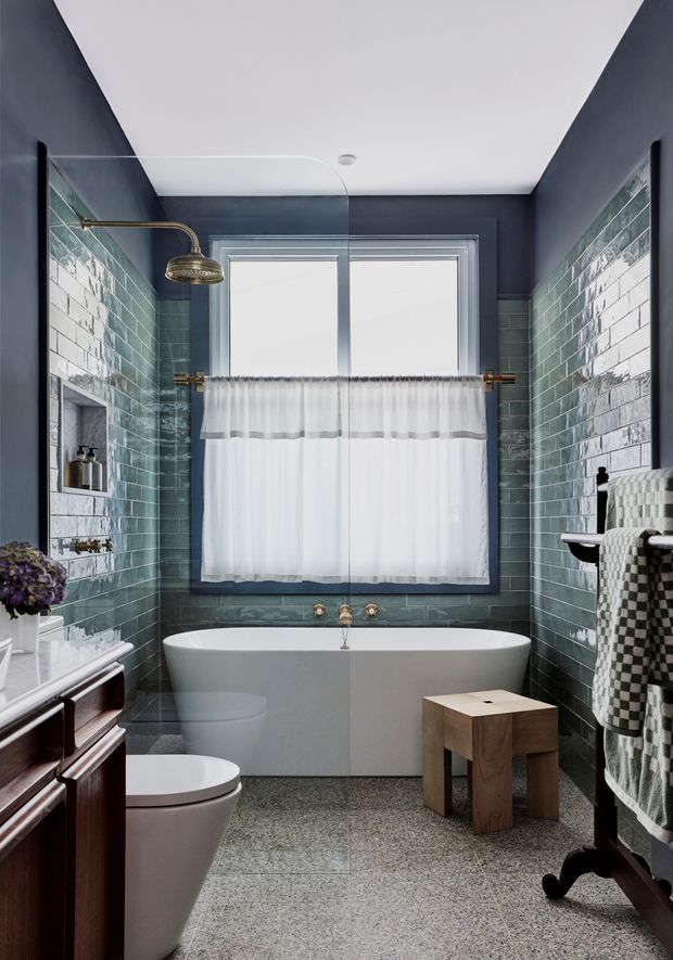 A compact, carefully detailed bathroom with a freestanding white bathtub beneath a curtained window, dark blue-grey painted upper walls, glossy green-grey subway tile wainscoting, and terrazzo-style flooring. A glass shower screen and brass shower fittings occupy the left side, while a dark timber vanity, toilet, towels, and a small wooden stool complete the room. The space has a refined traditional character with layered vintage-inspired details.