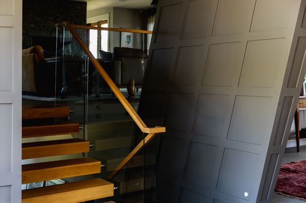 Interior view of a contemporary stairway with floating timber treads, clear glass balustrades, and a warm timber handrail. A large angled wall or partition clad in dark grey decorative rectangular panelling dominates the right side, with a darker living area visible beyond the stairs.