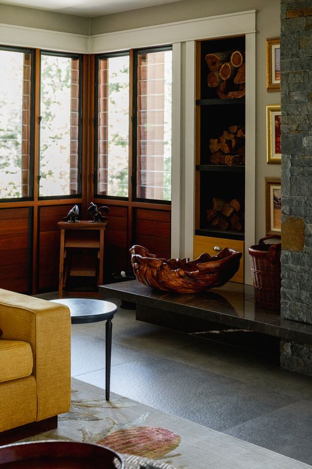 A warmly styled living room with tall corner windows framed in dark timber, white decorative trim, and wood-panelled lower walls. A yellow upholstered sofa, small round side table, carved timber bowl on a dark stone hearth ledge, built-in display shelving, framed artwork, woven basket, and a substantial grey stone-clad fireplace create a refined, nature-inspired interior.