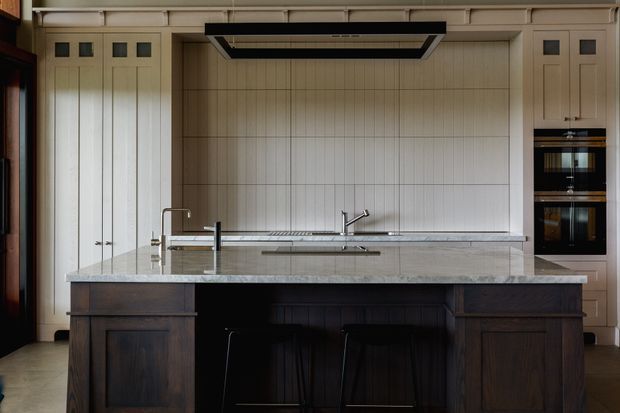 A refined traditional kitchen with a large dark timber island, pale painted cabinetry, integrated double ovens, a wide stone benchtop, and a full-height vertically panelled splashback. A sculptural black rangehood and undermount sink fixtures provide contemporary contrast, while the concealed appliances and symmetrical cabinetry create a tailored, high-end composition.