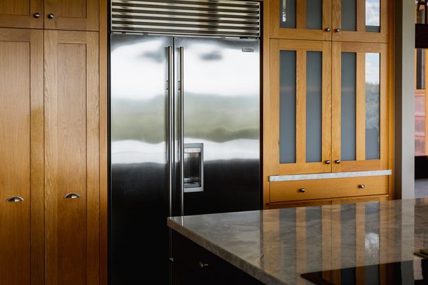 Close-up view of a refined kitchen featuring warm timber cabinetry, a large stainless-steel French-door refrigerator, integrated upper ventilation, and a polished stone island benchtop. The cabinetry includes framed glass-front doors and detailed hardware, creating a high-quality custom joinery aesthetic.