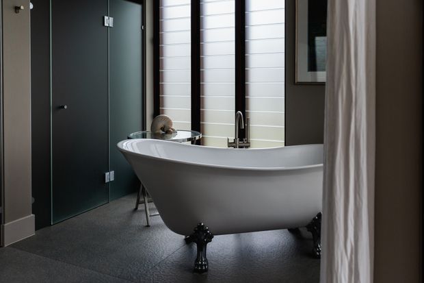 A moody, refined bathroom featuring a freestanding white clawfoot bathtub with dark ornate feet, a chrome floor-mounted tub filler, dark charcoal doors, white horizontal wall panelling, grey flooring, and a partially visible curtain and framed artwork.