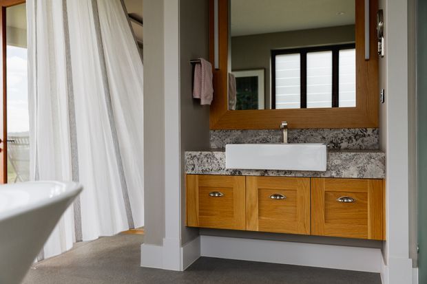 A contemporary bathroom with a floating timber vanity featuring three drawers, a white rectangular vessel basin, and a speckled stone benchtop and backsplash. A large timber-framed mirror sits above the vanity, while a freestanding white bathtub and sheer curtain are visible to the left. The room has neutral grey walls, carpeted flooring, and a bright, refined feel.