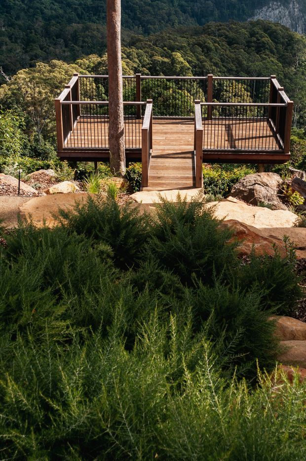 A timber viewing deck projects over a rugged, densely vegetated hillside, with dark metal balustrades and a short timber access bridge. Large rocks and native-looking planting occupy the foreground, while forested mountains and exposed cliff faces form the background. The scene is an outdoor landscape setting with a strong connection to nature.
