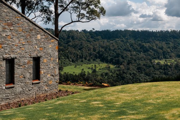 Exterior view of a rustic stone building set beside a broad manicured lawn, overlooking a heavily wooded rolling landscape. The facade features rough grey stonework with scattered warm ochre accents, narrow dark-framed windows, and a simple pitched roof edge.