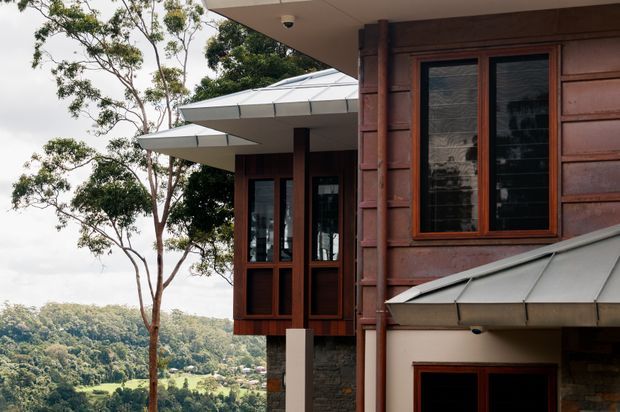 Exterior view of a modern multi-level house set into a leafy, sloping landscape. The facade combines dark stained timber cladding, stonework, extensive timber-framed glazing, projecting box-like window bays, and low-pitched metal roof planes with broad eaves.