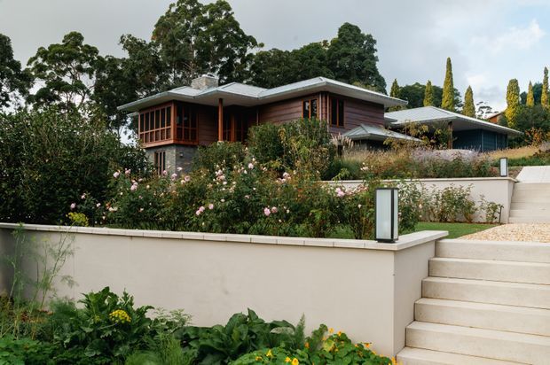 Exterior view of a low-profile, single-storey home set within a landscaped garden. The house features broad shallow roof planes, extensive timber-framed glazing, warm timber wall cladding, and a dark stone-clad base. Pale rendered retaining walls, broad steps, and flowering garden beds form the foreground.