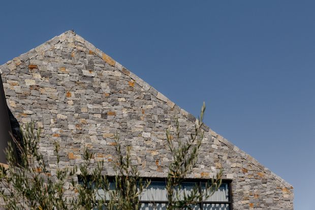 Exterior architectural detail showing a steep triangular gable wall constructed from irregular grey stone masonry with scattered warm ochre tones. A dark-framed glazed opening is partially visible at the base, screened by foreground vegetation, against a clear blue sky.
