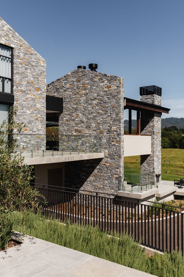 Exterior view of a contemporary multi-level residence built with extensive rough stone masonry, dark metal-framed glazing, and concrete terraces. The home steps down a sloping site, with glass balustrades, black metal railings, a flat roofline, and a chimney-like stone volume capped with dark horizontal screens. Rural hills and open landscape are visible beyond the property.