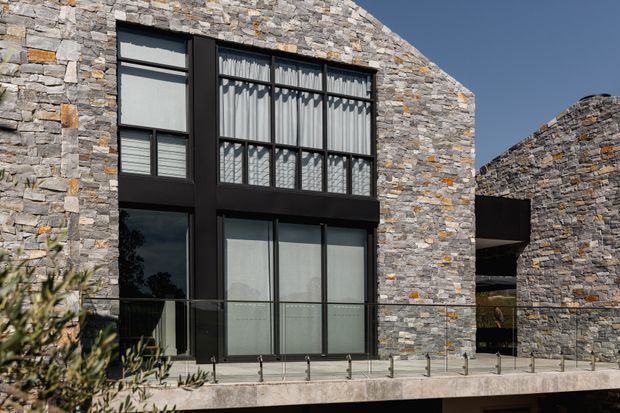 Contemporary exterior facade featuring extensive irregular natural stone cladding in grey, beige and rust tones, large black-framed windows, and a projecting glazed balcony with clear glass balustrading. The building has curved or organically shaped stone walls and a strong connection between solid masonry and expansive glazing.