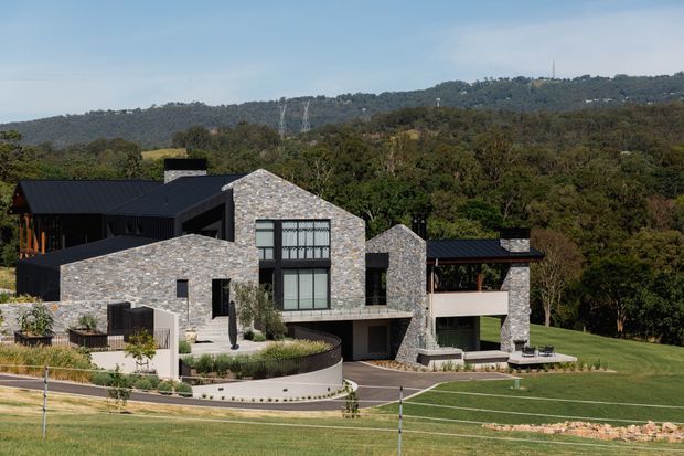 A large contemporary multi-level residence set into a landscaped, wooded hillside. The building features irregular natural-stone cladding, dark metal pitched and flat roof forms, expansive black-framed glazing, terraces, retaining walls, and a broad manicured lawn. Utility transmission lines are visible on the distant ridge.
