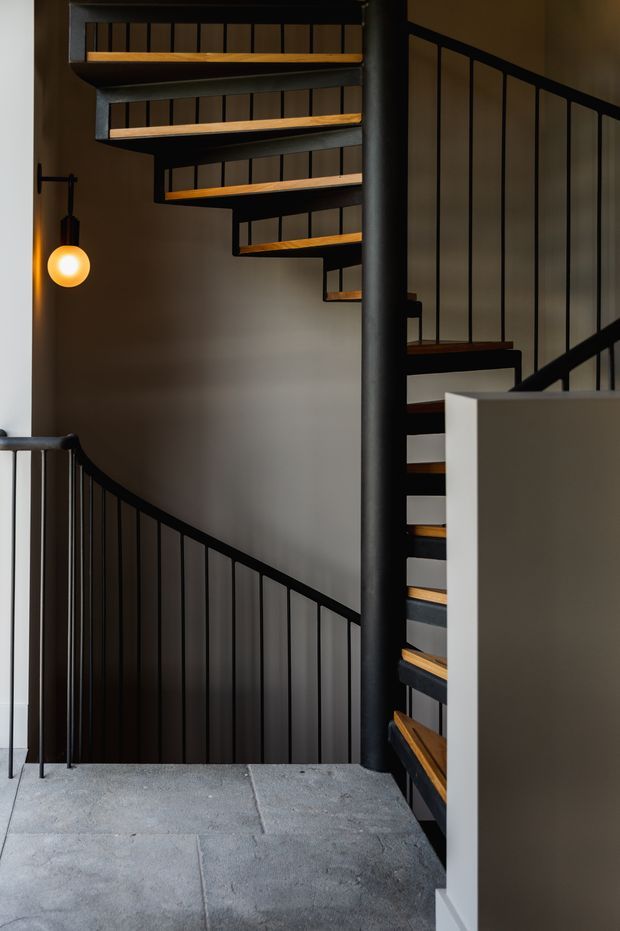 A dramatic interior view of a multi-level spiral staircase with black metal railings and central post, warm timber stair treads, grey concrete flooring, and dark neutral walls. A small spherical pendant light adds a warm glow beside the stairwell.