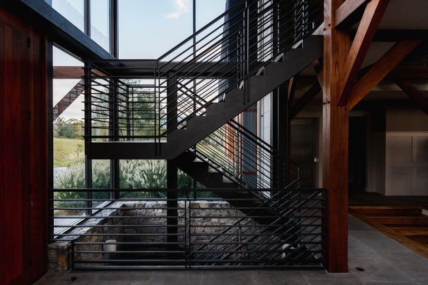 A dramatic interior stair hall with a multi-level black metal staircase and fine horizontal railings set against expansive floor-to-ceiling glazing. Exposed dark timber posts and beams frame the space, with stone landscaping visible beyond the glass and a polished dark floor underfoot. The composition emphasizes strong structural lines, transparency, and a connection to the surrounding landscape.