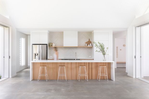 A bright, open-plan contemporary kitchen viewed straight on. The room features a large island with vertical timber slat detailing, a pale stone or engineered-stone benchtop, white cabinetry, a stainless-steel refrigerator, a light stone splashback, and four slim bar stools. Large glazed openings and pale grey polished concrete flooring create an airy, minimal setting.