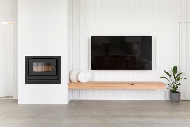 Bright minimalist living room featuring a black built-in fireplace, wall-mounted television, white vertical wall panelling, and a long light timber floating media shelf. A potted plant and simple sculptural decor add restrained natural accents, while the polished concrete floor contributes an understated contemporary feel.