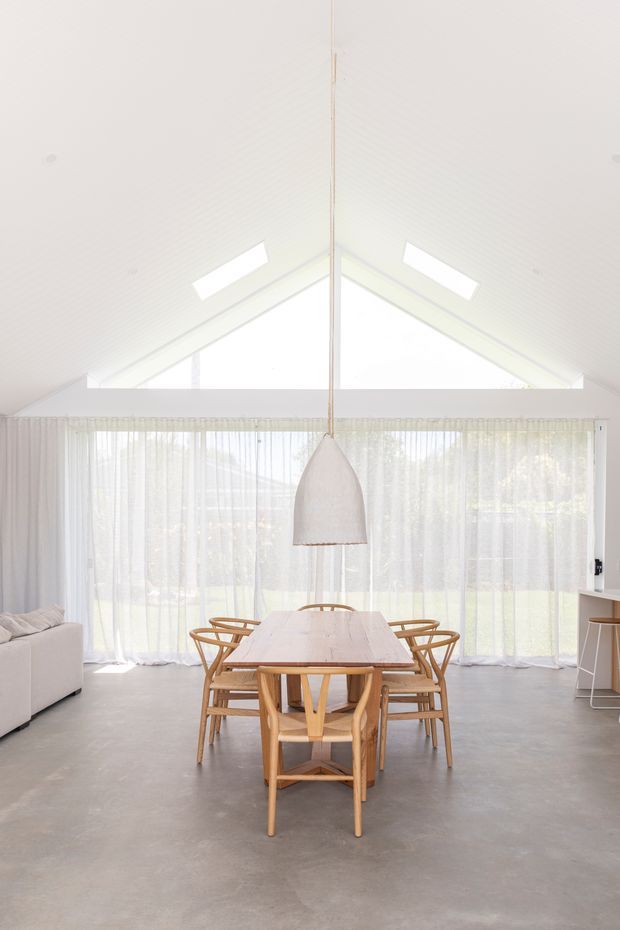 Bright, airy open-plan dining and living space with a long light-wood dining table surrounded by timber chairs, a large sculptural pendant light, sheer floor-to-ceiling curtains, and expansive glazing. The room has a high vaulted white ceiling with skylights and a triangular clerestory window, polished grey concrete flooring, and minimalist white built-in cabinetry.