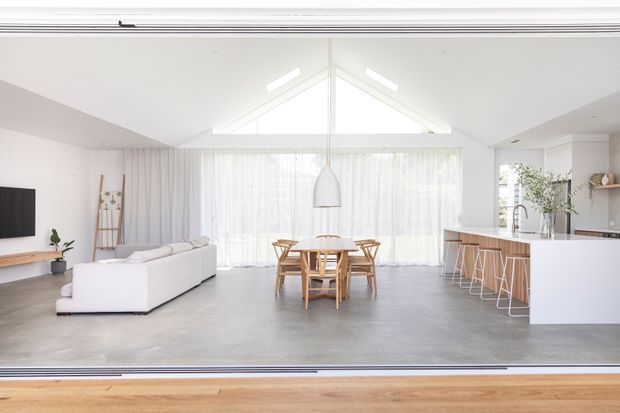 Bright open-plan interior with a minimalist living, dining and kitchen arrangement. The room features a white pitched ceiling with clerestory-style glazing, sheer curtains, polished grey concrete flooring, a pale upholstered sofa, light timber dining furniture, and a white kitchen island with bar seating. Large glazed openings create an airy connection to the outdoors.