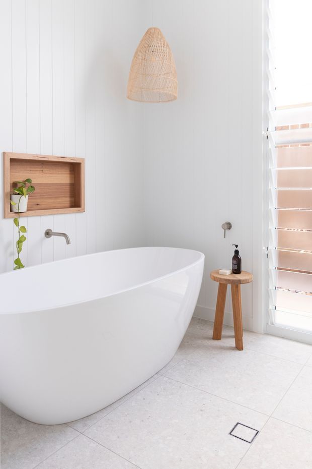 Bright, serene bathroom featuring a freestanding white bathtub, white vertical wall panelling, pale stone-look floor tiles, a timber recessed display niche, wall-mounted tapware, a woven pendant light, and louvered glazing. A small timber stool with toiletries and a potted plant add natural warmth.