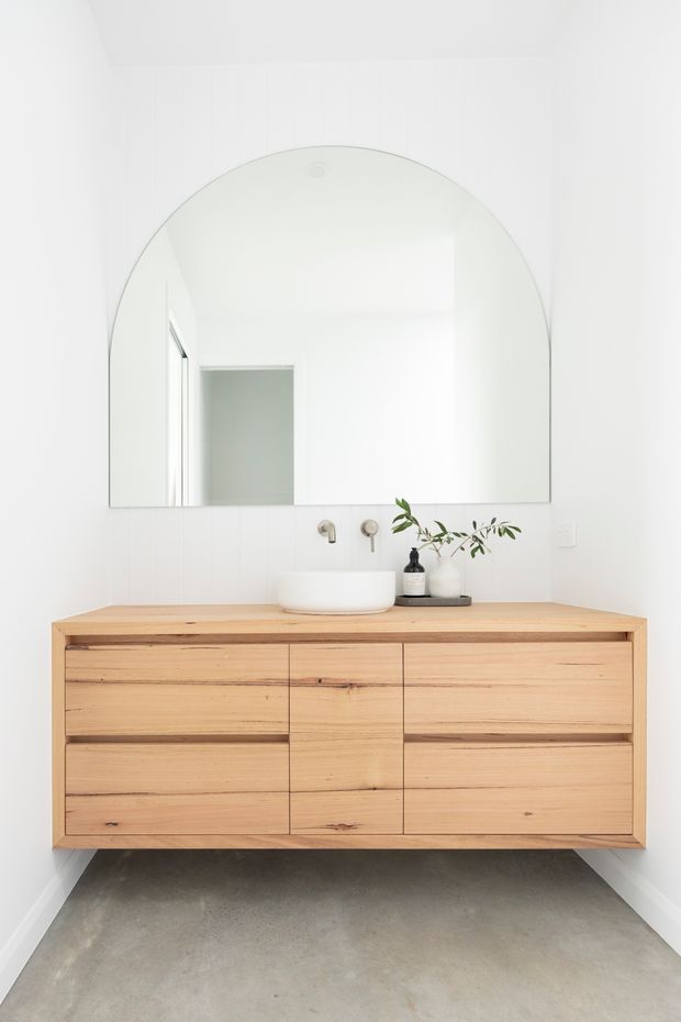 Bright minimalist bathroom featuring a wall-mounted natural timber vanity with integrated drawers, a white vessel basin, wall-mounted tapware, and a large arched mirror. White vertical wall tiles and a smooth grey concrete-look floor create a clean, contemporary appearance.