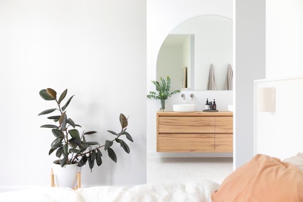 Bright, minimalist bedroom scene with a partially visible bed in the foreground, looking toward an open bathroom vanity area. The bathroom features a floating timber vanity with a white vessel basin, wall-mounted tapware, an arched mirror, and soft grey-white finishes. A potted rubber plant adds natural greenery, while peach bedding provides a warm accent.