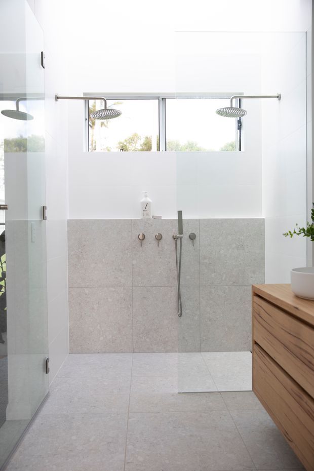 Bright contemporary bathroom featuring a large frameless glass walk-in shower with dual rain showerheads, pale stone-look wall and floor tiles, white upper walls, and a timber vanity with integrated drawers. A high horizontal window provides natural light and views of greenery.