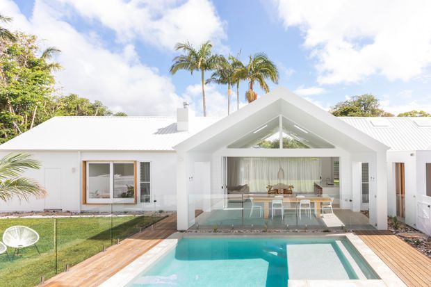 Bright tropical residential exterior viewed from the pool deck, featuring a white single-storey home with a pitched gable entry pavilion, white metal roof, extensive glazing, and a covered outdoor dining area overlooking a turquoise swimming pool. Timber decking, glass pool fencing, palms, and lush landscaping reinforce the resort-like setting.