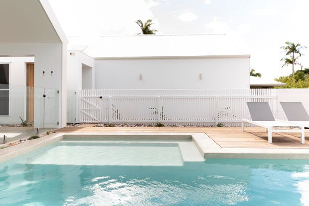 Bright contemporary white residential exterior with a swimming pool in the foreground, timber pool deck, white vertical-slat fencing, flat-roofed or low-pitched forms, and tropical palms in the background. The scene has a clean coastal resort-like feel with strong white and aqua tones.