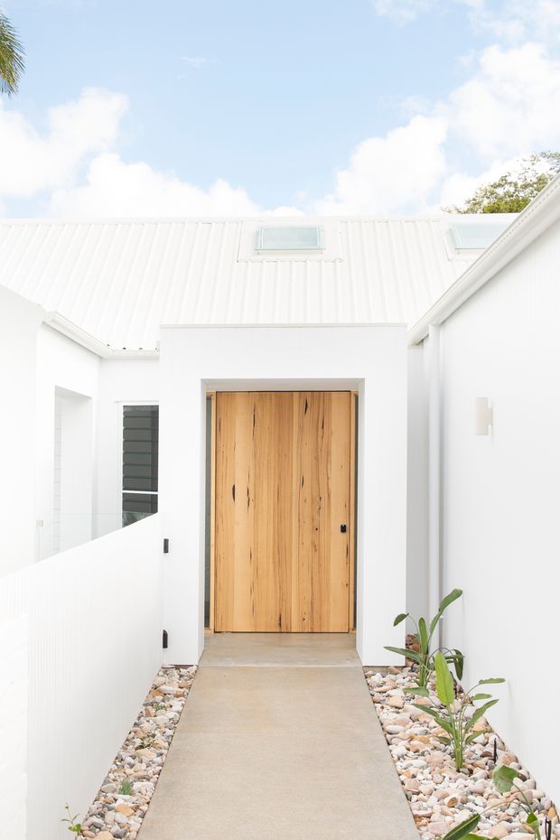 Bright minimalist residential entry courtyard with a large natural timber front door set into a white rendered facade. A white ribbed metal roof with skylights is visible above, while a narrow concrete pathway is bordered by decorative stones and sparse tropical planting.