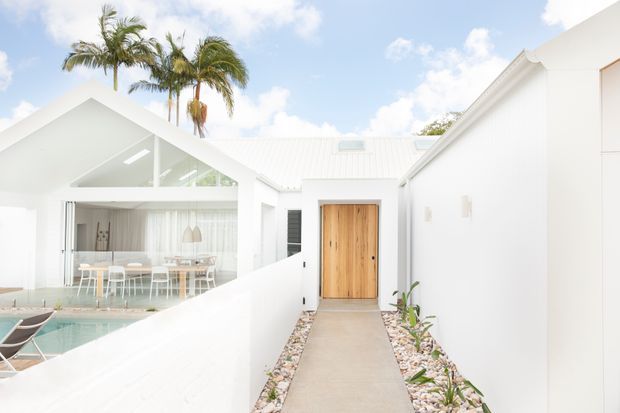 Bright white single-storey residential exterior with gabled roof forms, a timber pivot entry door, and a concrete path bordered by decorative gravel and planting. A glazed dining area and swimming pool are visible within the courtyard, with tall palms behind the home.