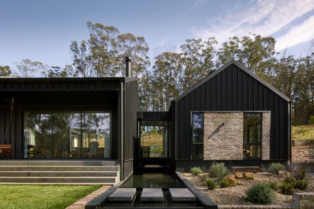 Exterior view of a contemporary single-storey residence composed of dark vertical-clad gabled and flat-roofed volumes. Large glazed living areas open toward a landscaped entry with a narrow reflecting-water feature and stepping stones. A light stone or brick feature wall contrasts with the dark facade, with native planting and mature trees surrounding the home.