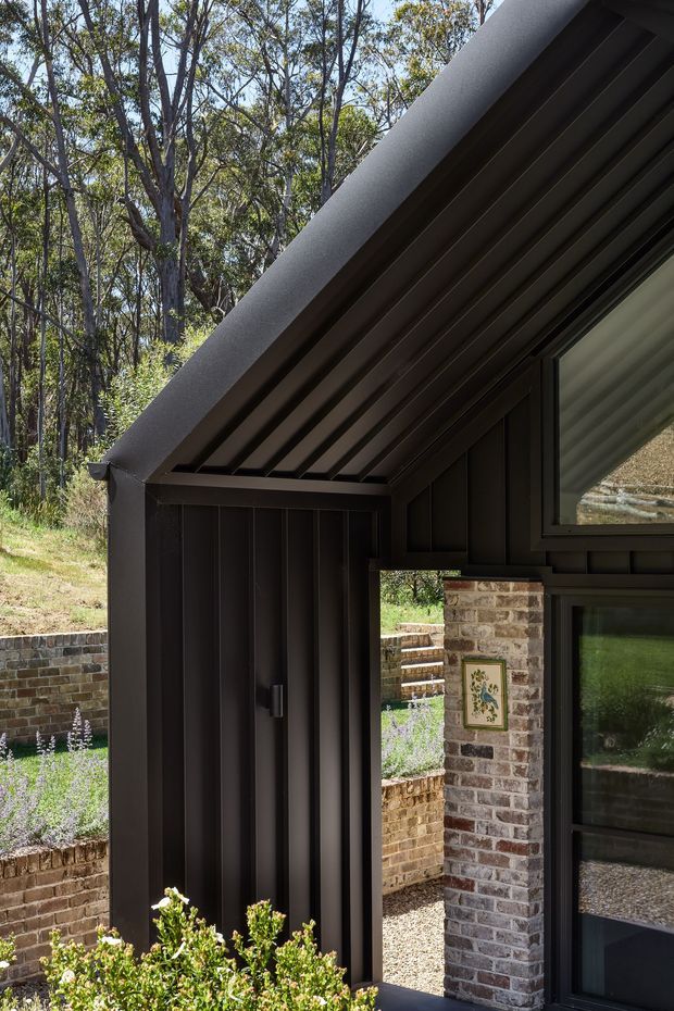 Close exterior architectural view of a dark-clad building entrance or covered passage in a wooded setting. The steep pitched roof has exposed dark metal rafters or lining, with dark vertical cladding, black-framed glazing, and a contrasting exposed brick wall and column. Terraced brick garden walls and planting are visible around the structure.