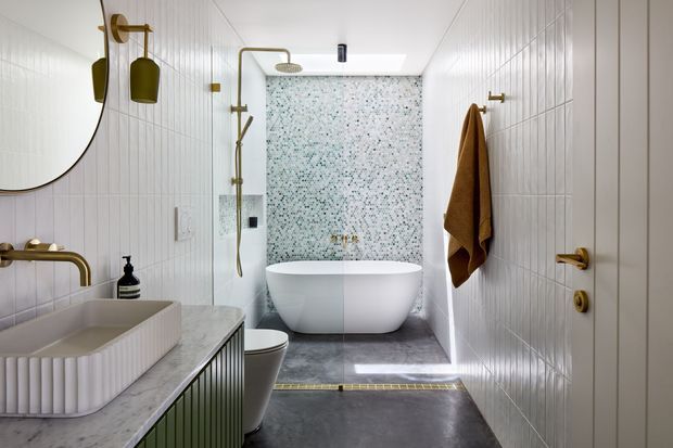 A compact, refined bathroom with white vertically tiled walls, a freestanding white bathtub, wall-mounted shower, patterned mosaic feature wall, and a sculptural ribbed vanity with a pale stone benchtop. Brass tapware, fittings, lighting, and towel hardware add warm metallic accents, while dark stone-look flooring creates contrast. A mirror, basin, toilet, and shower zone are visible.