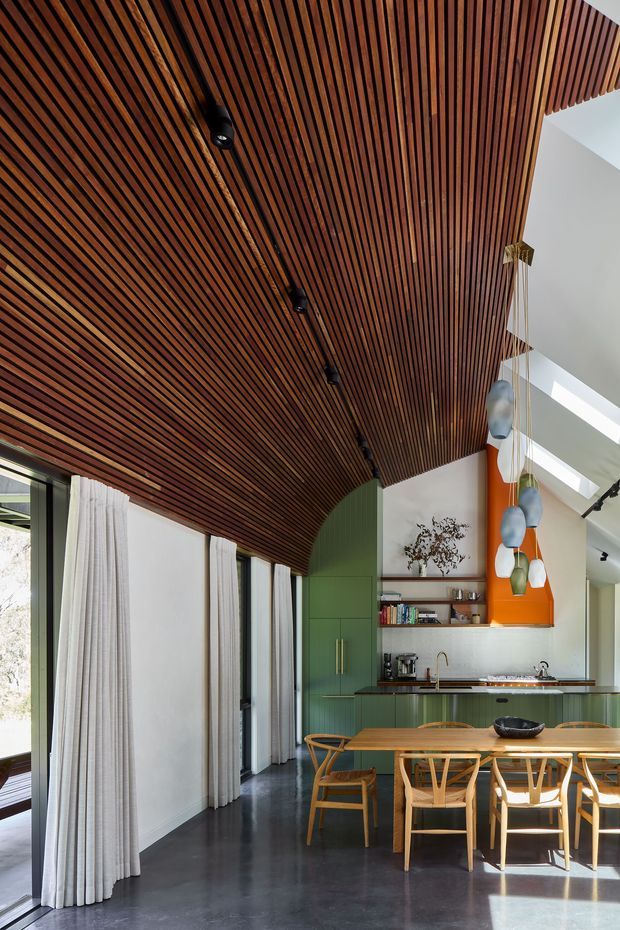 A bright, double-height open-plan kitchen and dining area with a dramatic curved ceiling clad in narrow dark timber battens. The space features green built-in kitchen cabinetry with brass-toned handles, a pale stone benchtop and splashback, a timber dining table, sculptural timber dining chairs, dark polished flooring, full-height curtains and large glazing. A vivid orange vertical feature wall and clustered pendant lights add colour and visual interest.