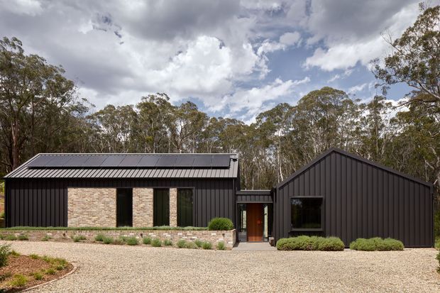 Exterior view of a contemporary single-storey home composed of two connected dark-clad volumes. The left wing has a low-pitched roof with solar panels, vertical dark metal cladding, pale brick feature walls, and tall narrow windows. The right wing has a prominent front-facing gable with matching vertical dark cladding and a large window. A glazed or enclosed link connects the volumes, opening onto a gravel forecourt with low landscaped planting, backed by mature woodland under a cloudy sky.