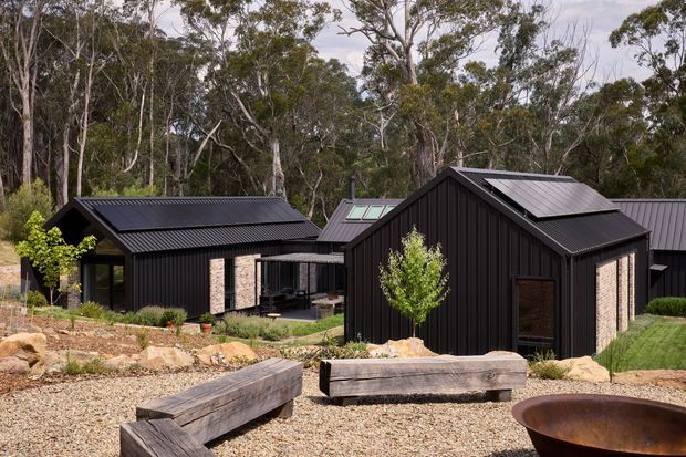 A contemporary cluster of dark-clad, single-storey buildings set within a native wooded landscape. The structures feature steep pitched roofs, black vertical metal cladding, pale masonry accent walls, extensive glazing, and rooftop solar panels. A gravelled landscaped courtyard in the foreground includes large timber benches and a rusted circular fire bowl.