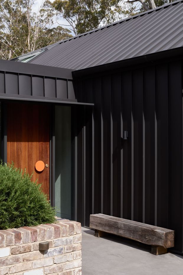 A close-up exterior view of a contemporary dark-clad building entry. The facade features vertically ribbed black metal cladding, a pitched corrugated metal roof, a warm timber entry door with a bright orange circular handle, and a simple heavy timber bench. A low curved brick planter with dense greenery frames the entrance.