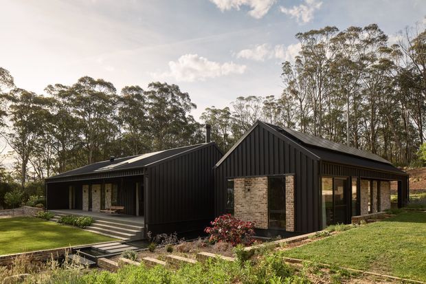A low, modern dark-clad house set within a landscaped, wooded site. The building is composed of connected gabled volumes with black vertical cladding, dark metal roofing, expansive glazed openings, and contrasting pale brick feature walls. Stepped paths, terraces, lawn, and garden beds surround the residence.