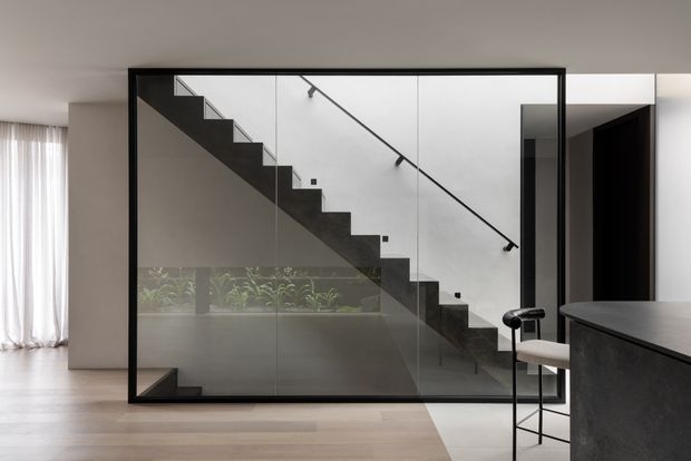 Contemporary interior featuring a minimalist black-framed glass enclosure around a straight staircase. The staircase has dark treads, a solid dark side profile, and a slim black handrail, with planting visible behind the glazing. Pale timber flooring, white walls, sheer curtains, and a dark kitchen island with a bar stool create a refined, monochromatic composition.