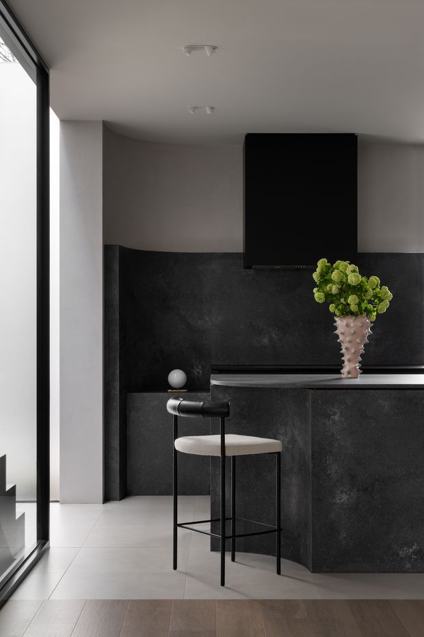 A minimalist contemporary kitchen or bar area with dark stone-look cabinetry and backsplash, a slim black countertop, and a single upholstered bar stool. A large black rectangular rangehood or wall panel dominates the rear wall, while floor-to-ceiling glazing brings in daylight along the left side. Neutral grey walls, pale large-format floor tiles, dark timber flooring in the foreground, and a sculptural vase with green flowers complete the refined monochrome interior.