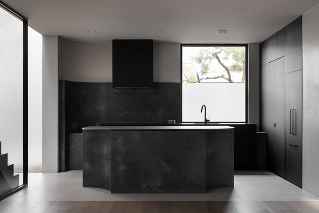 A sleek, contemporary kitchen dominated by a large dark island with an integrated cooktop and overhead rangehood. Matching dark cabinetry lines the rear and right walls, with a black splashback, undermount sink, and minimalist black tap beneath a large window. Pale tiled flooring and light walls provide contrast, while full-height glazing at left brings in natural light.