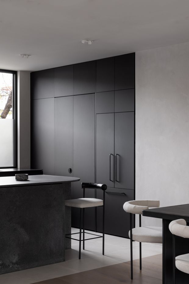 A refined monochromatic kitchen and dining area with a large matte-black built-in cabinetry wall, integrated tall storage, dark stone island, pale smooth walls, and minimalist black-framed window. A small dining table with upholstered chairs occupies the foreground, while slim black bar stools sit at the island. The space has clean linear detailing, restrained contemporary furnishings, and soft natural light.