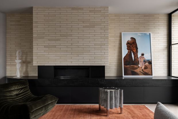 Contemporary living room with a black built-in fireplace and low cabinetry set against a light exposed-brick feature wall. A dark green sculptural sofa, copper-toned rug, small metallic coffee table, framed artwork, and expansive black-framed glazing create a refined, warm-toned interior.