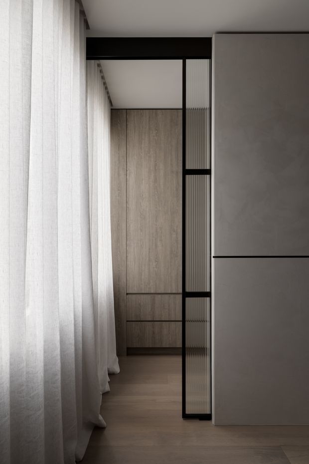 A quiet, minimalist interior view featuring full-height sheer white curtains along the left side, warm timber flooring and matching timber-toned built-in cabinetry at the rear. A slim black-framed ribbed glass partition creates a screened transition, while a large smooth grey built-in wall or cupboard occupies the right side. The composition is vertically layered, softly lit, and highly restrained.