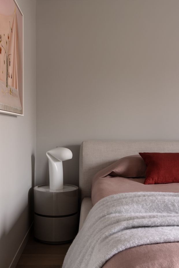 A close-up view of a refined bedroom corner featuring a softly upholstered taupe bedhead, dusty rose bedding, a deep red accent cushion, and a round dark bedside table with a sculptural white lamp. The walls are painted warm grey-beige, with framed botanical artwork partially visible on the left and dark timber flooring below. The styling is restrained, warm, and contemporary.