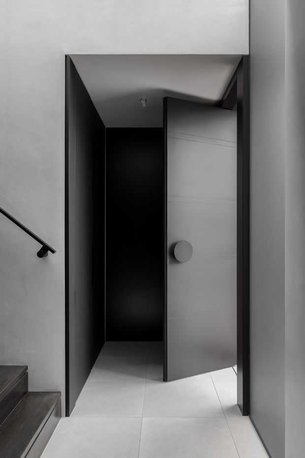 A monochromatic minimalist interior view of a narrow doorway or passage beneath a sloping ceiling. The space features dark charcoal door frames and walls, a partially open light grey door with a round knob, pale large-format tiled flooring, smooth light plaster walls, and a simple black stair handrail visible at left. The stark contrast and concealed, angular entry create a refined, architectural feel.