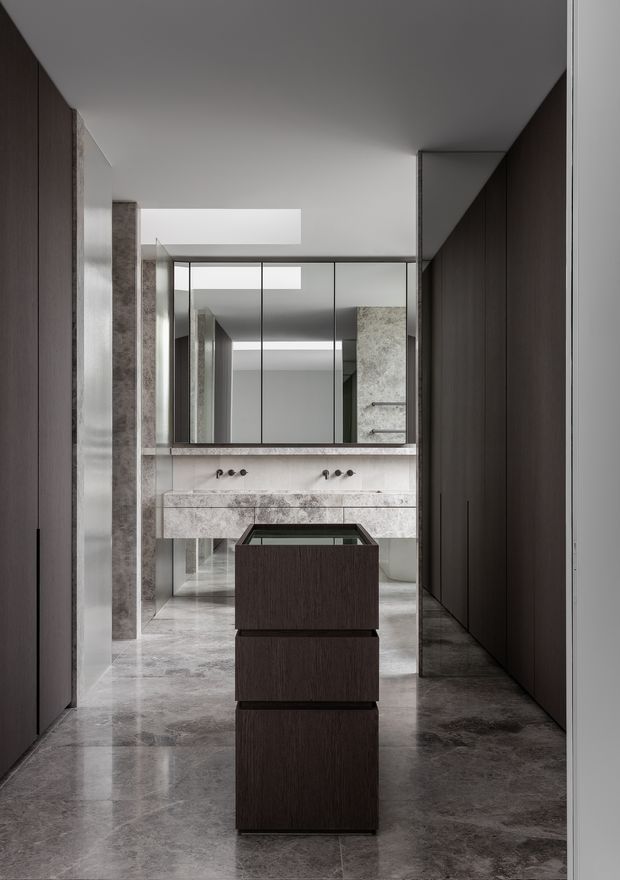 A refined, minimalist bathroom viewed through a dark timber-lined corridor. The space features a long stone vanity with twin basins and wall-mounted taps, large mirrored cabinetry, pale stone wall surfaces, polished grey stone flooring, and a central dark timber island or storage unit. Strong axial symmetry and reflective surfaces create a calm, luxurious atmosphere.