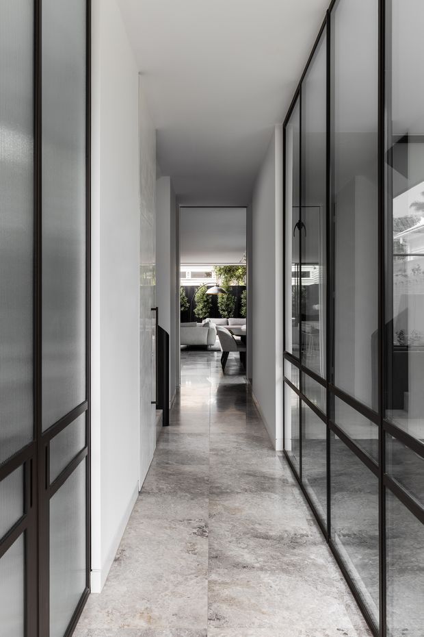 A long, narrow contemporary hallway with white walls, large-format grey stone flooring, and black-framed glass partitions and doors. The corridor leads toward a bright living area with a sofa and greenery visible at the end, while the glazing reflects the hall and adjacent spaces.