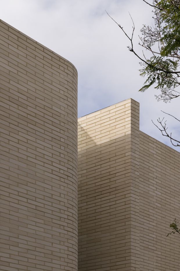 Close-up exterior architectural view of a contemporary building composed of pale cream masonry brick walls. The image emphasizes a tall curved brick facade meeting rectilinear brick volumes, with clean geometric lines and minimal detailing. Sparse tree branches frame the upper right against an overcast sky.