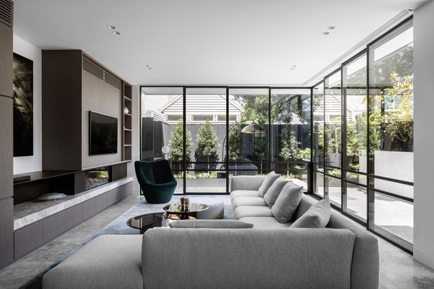 A bright contemporary living room with a large light-grey sectional sofa, dark green accent chair, round coffee table, and built-in media cabinetry. Floor-to-ceiling black-framed glazing wraps the room, opening views to a landscaped courtyard and bringing abundant natural light into the space. The interior has a refined neutral palette with dark timber joinery and polished concrete or stone flooring.