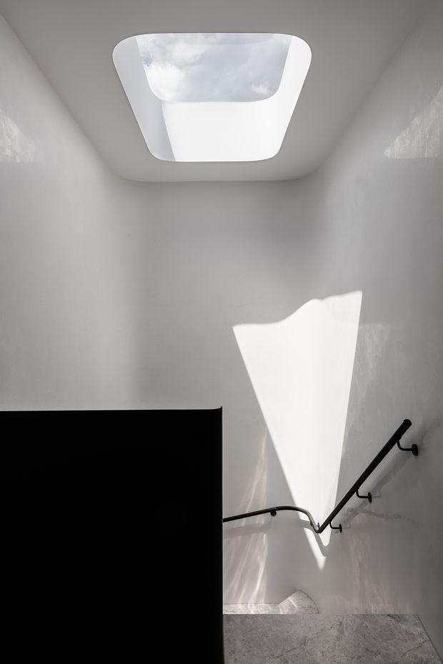 A minimalist interior stairwell with white plaster walls, a sculptural skylight, and strong natural light casting dramatic geometric shadows. A black metal handrail runs along the stair, contrasting with the pale textured floor and walls.